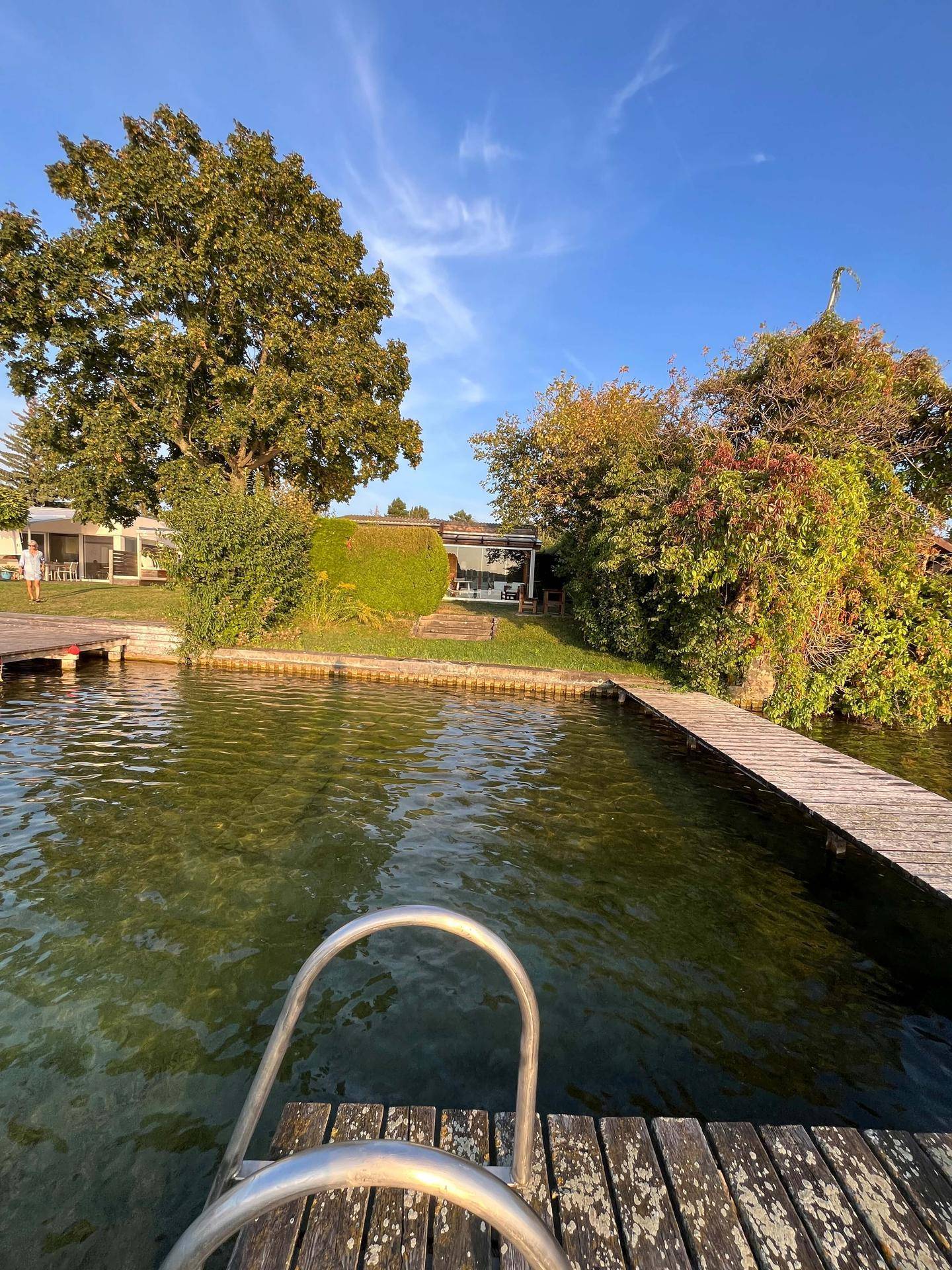Blick vom Wasser auf den Badesteg mit Leiter und das dahinterliegende Holzhaus am Seeufer.