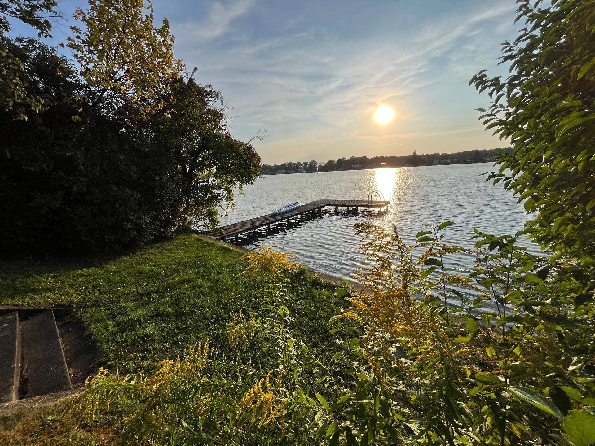 Malerischer Seeblick mit Holzsteg und üppiger Ufervegetation im warmen Abendlicht.