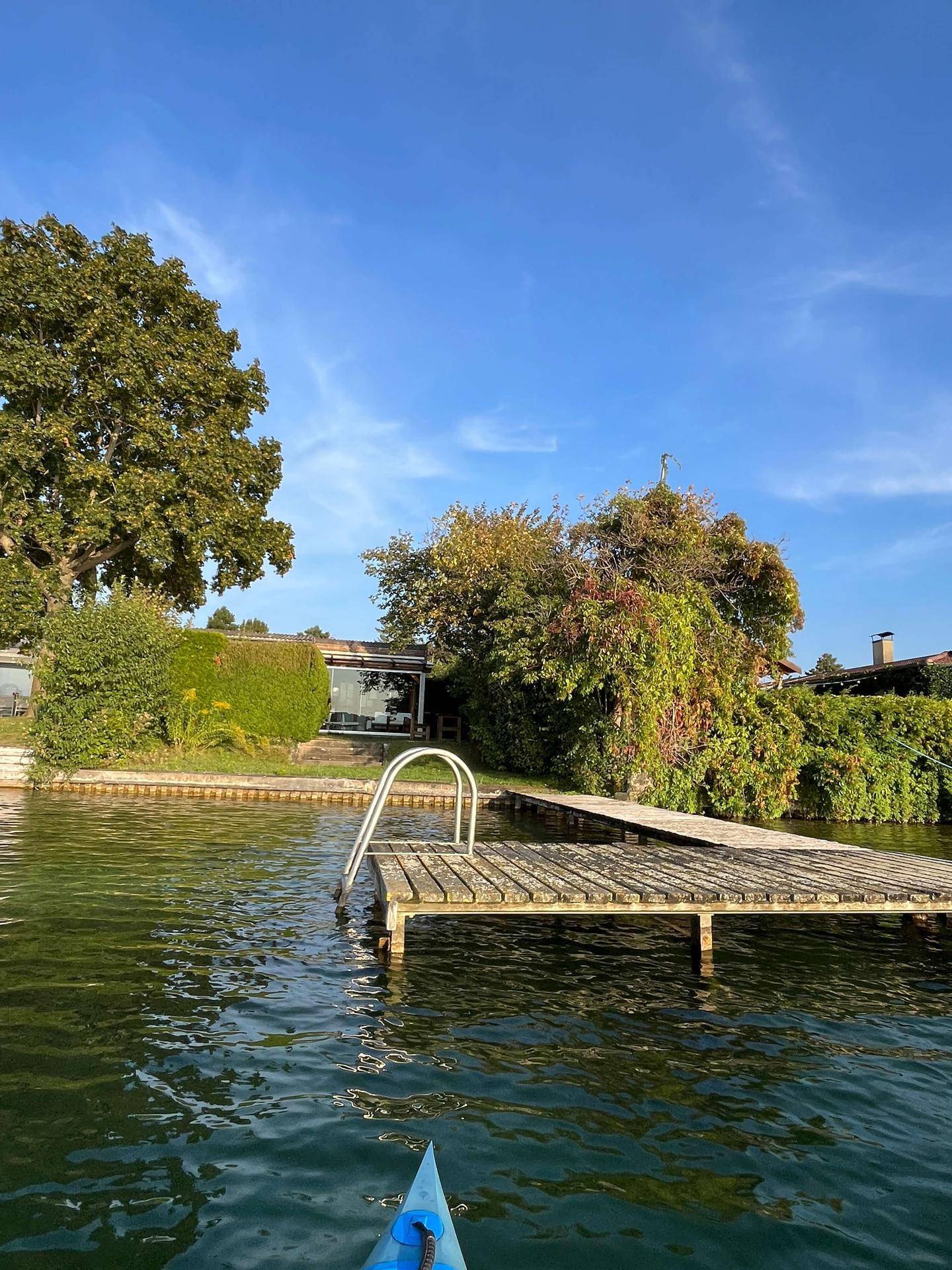 Perspektive vom Kajak auf den See, den Steg und das charmante Holzhaus am Ufer.