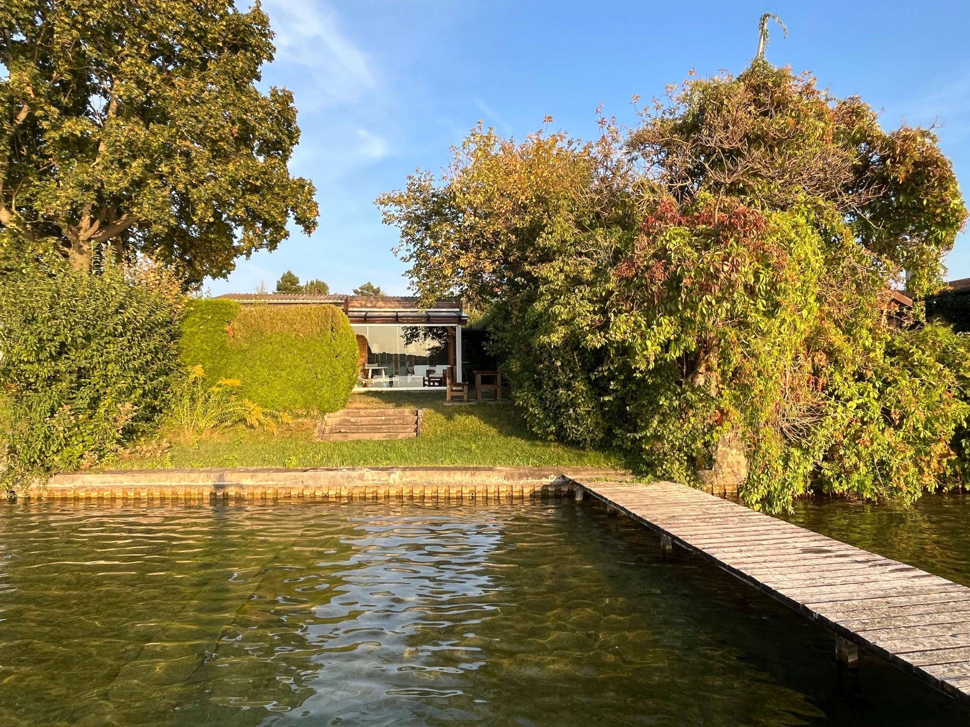 Blick vom See auf das charmante Holzhaus mit eigenem Steg und grüner Uferzone.