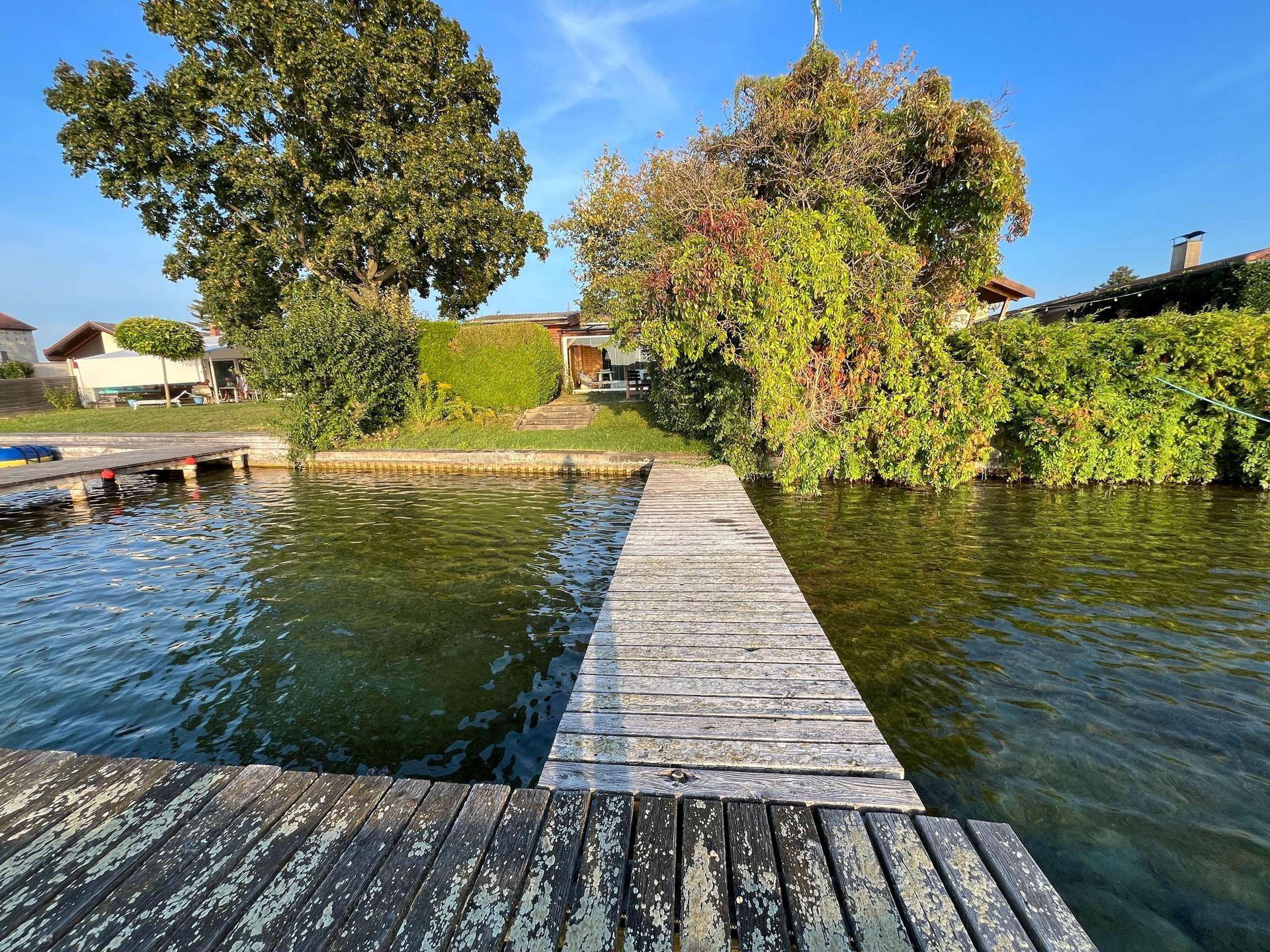 Weitläufiger Blick vom See auf das Holzhaus mit Steg und die umliegende Seenlandschaft.