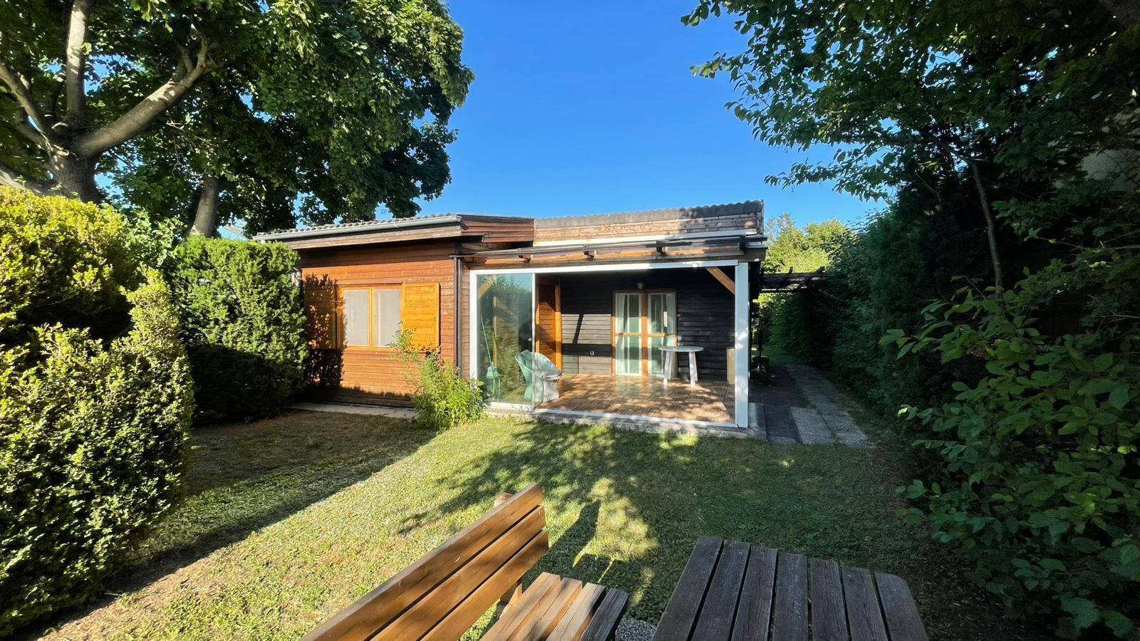 Gemütliches Holzhaus mit Gartenbereich und rustikaler Holzbank unter schattenspendenden Bäumen.