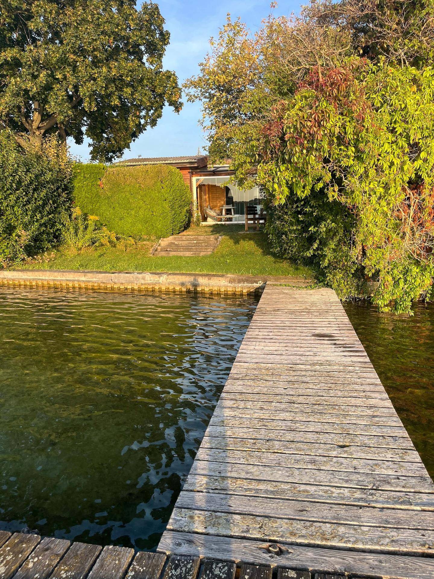 Idyllischer Blick vom Wasser auf das Holzhaus mit privatem Steg und üppiger Vegetation am Ufer.