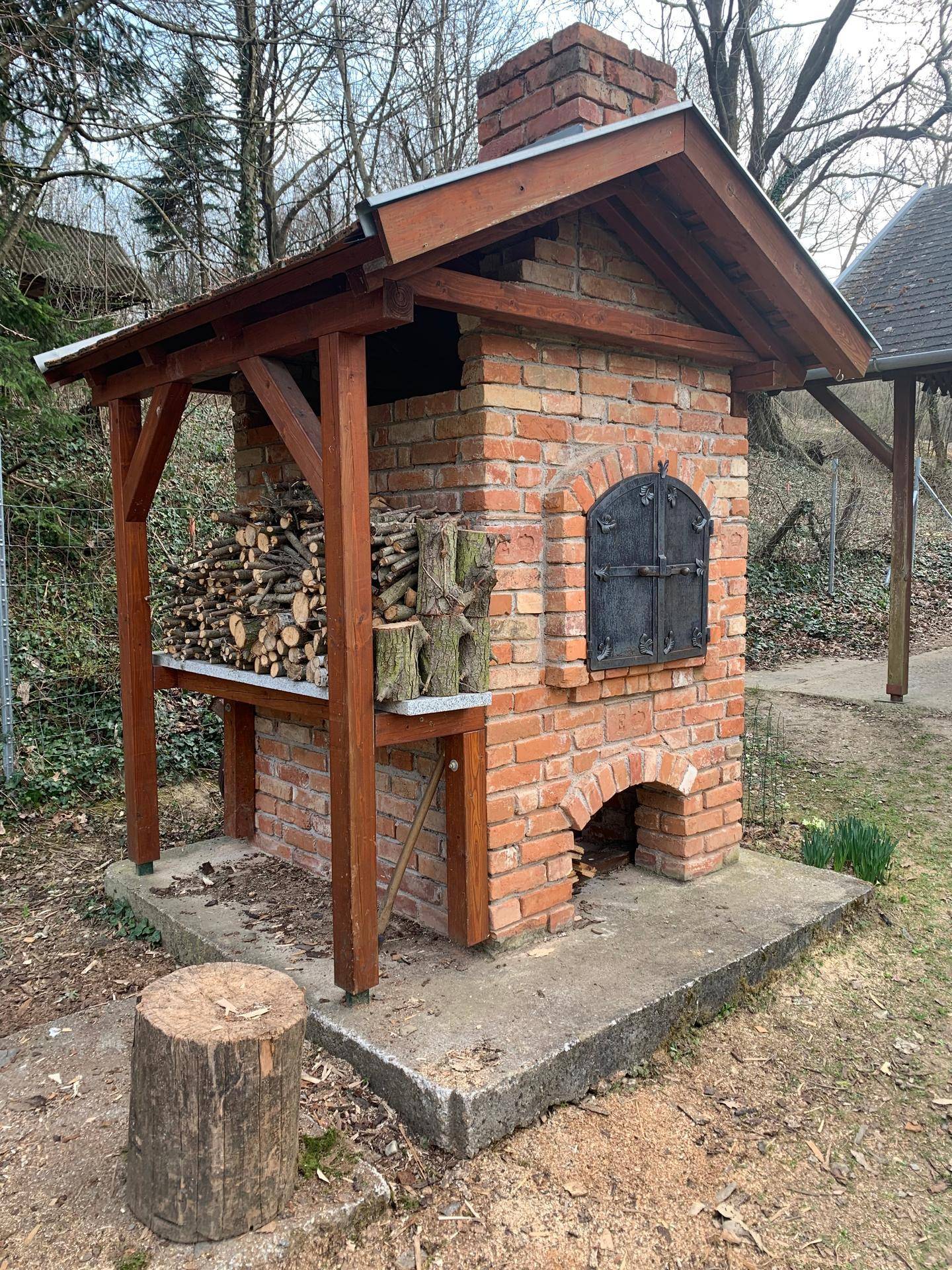 Traditioneller gemauerter Backofen im Freien mit Holzunterstand und gestapeltem Brennholz.