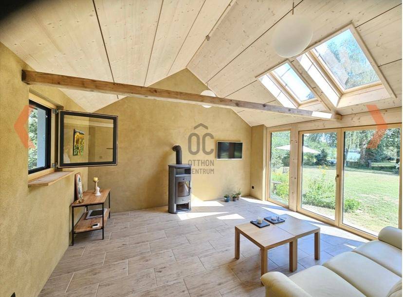 Helles Wohnzimmer mit Holzbalkendecke, Kaminofen und großen Fenstern mit Gartenblick.