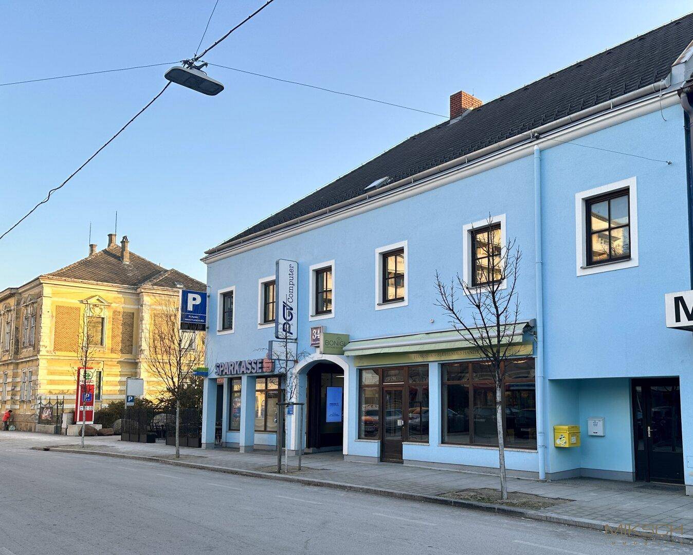 Ansicht eines hellblauen Geschäftsgebäudes an einer Straße mit großen Fenstern und umliegender Bebauung.
