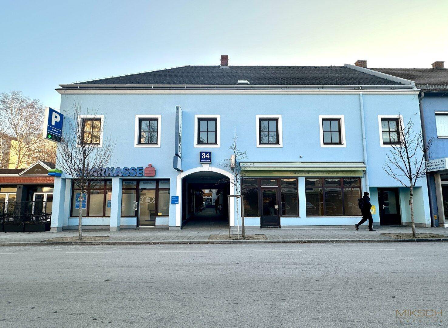 Breite Fassade eines hellblauen Geschäftsgebäudes mit mehreren Ladenlokalen und einem zentralen Durchgang.