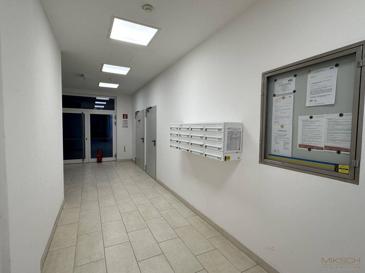 Langer Flur mit Briefkästen, einer Informationstafel und hellen Fliesen auf dem Boden.