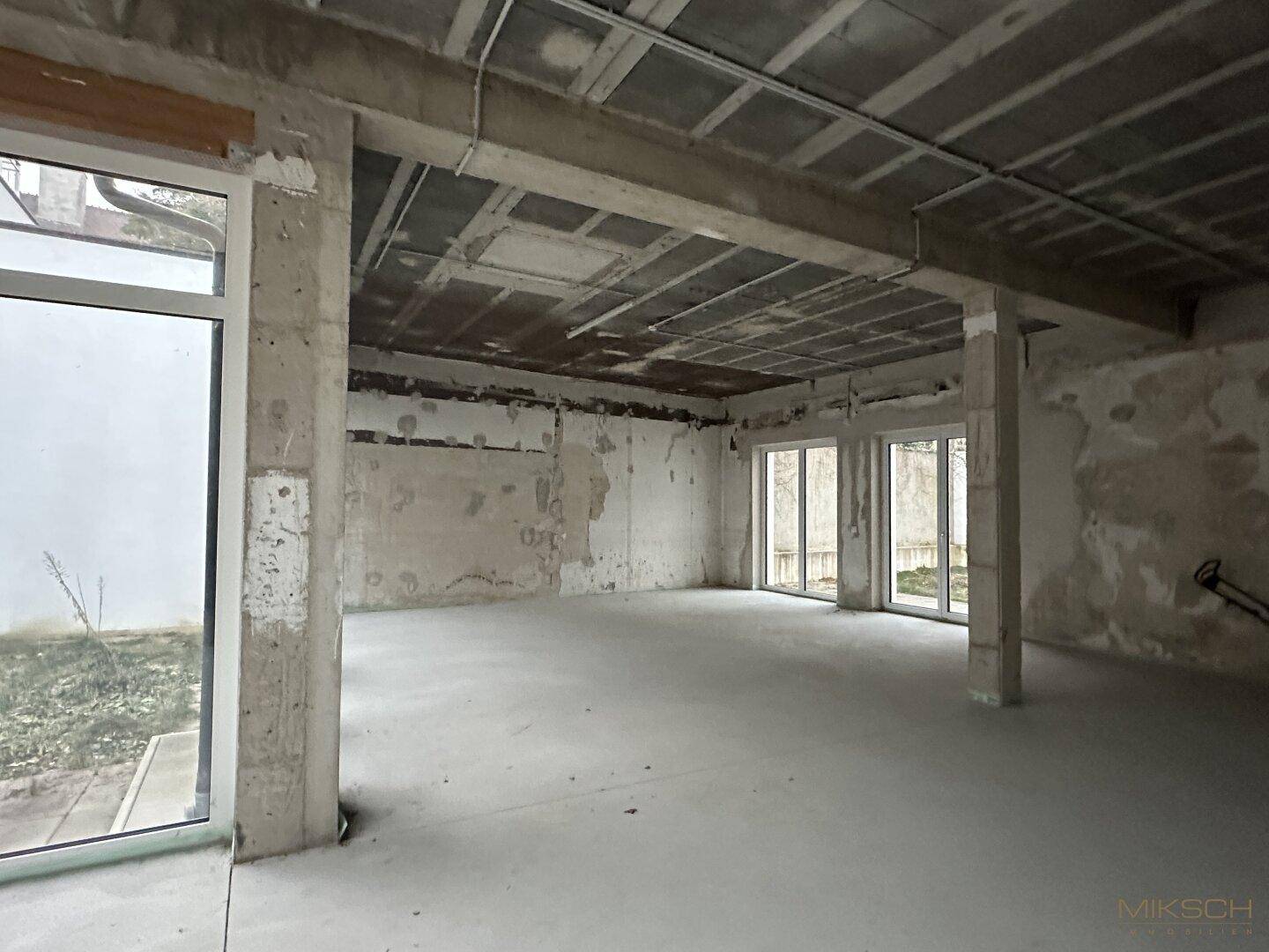 Weiterer Blick in den großen, unfertigen Gewerberaum mit Betonstrukturen und Fenstern zum Außenbereich.