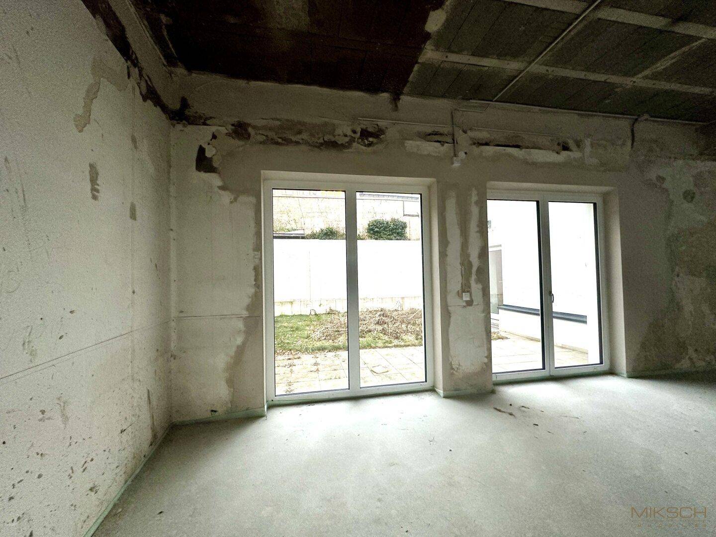 Ecke des unfertigen Innenraums mit großen Fenstern, die auf einen kleinen, ungepflegten Außenbereich blicken.