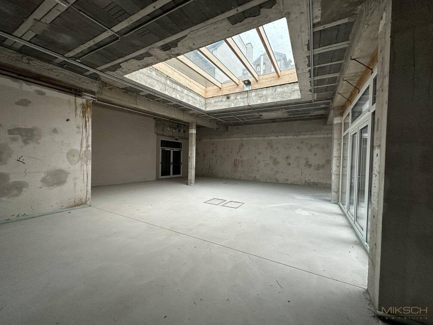 Weitwinkelaufnahme des unfertigen Gewerberaums mit Betonwänden, hohen Decken und einem großen Dachfenster.