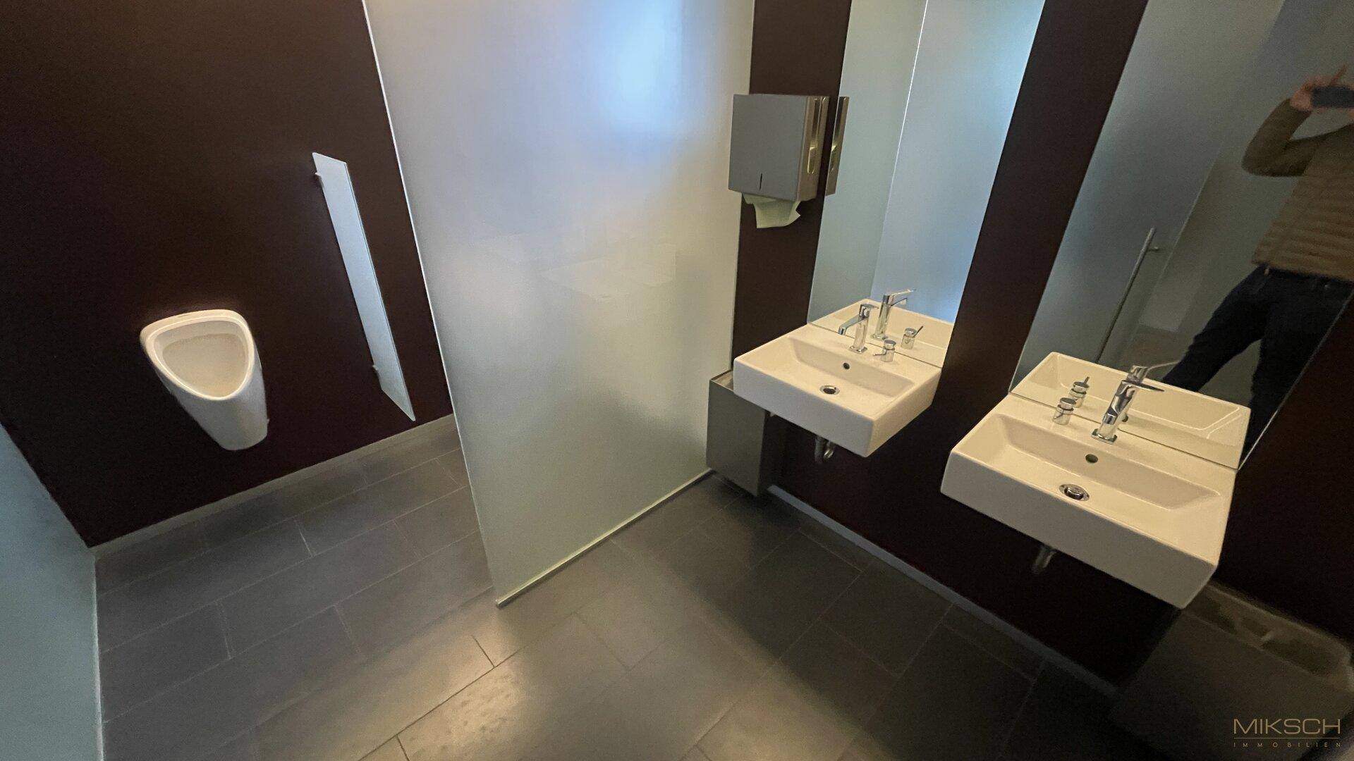 Moderne Herrentoilette mit Urinal, zwei Waschbecken und dunklen Fliesen.