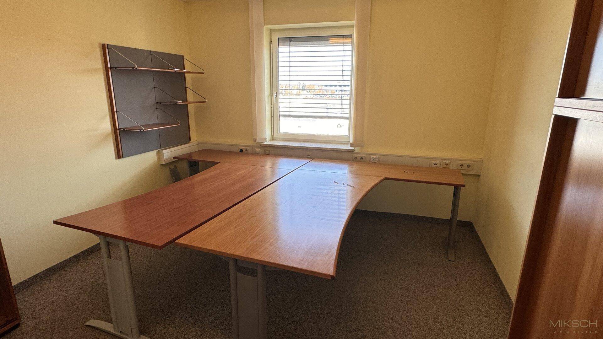 Kleines Büro mit gelben Wänden, Teppichboden und einem L-förmigen Schreibtisch am Fenster.
