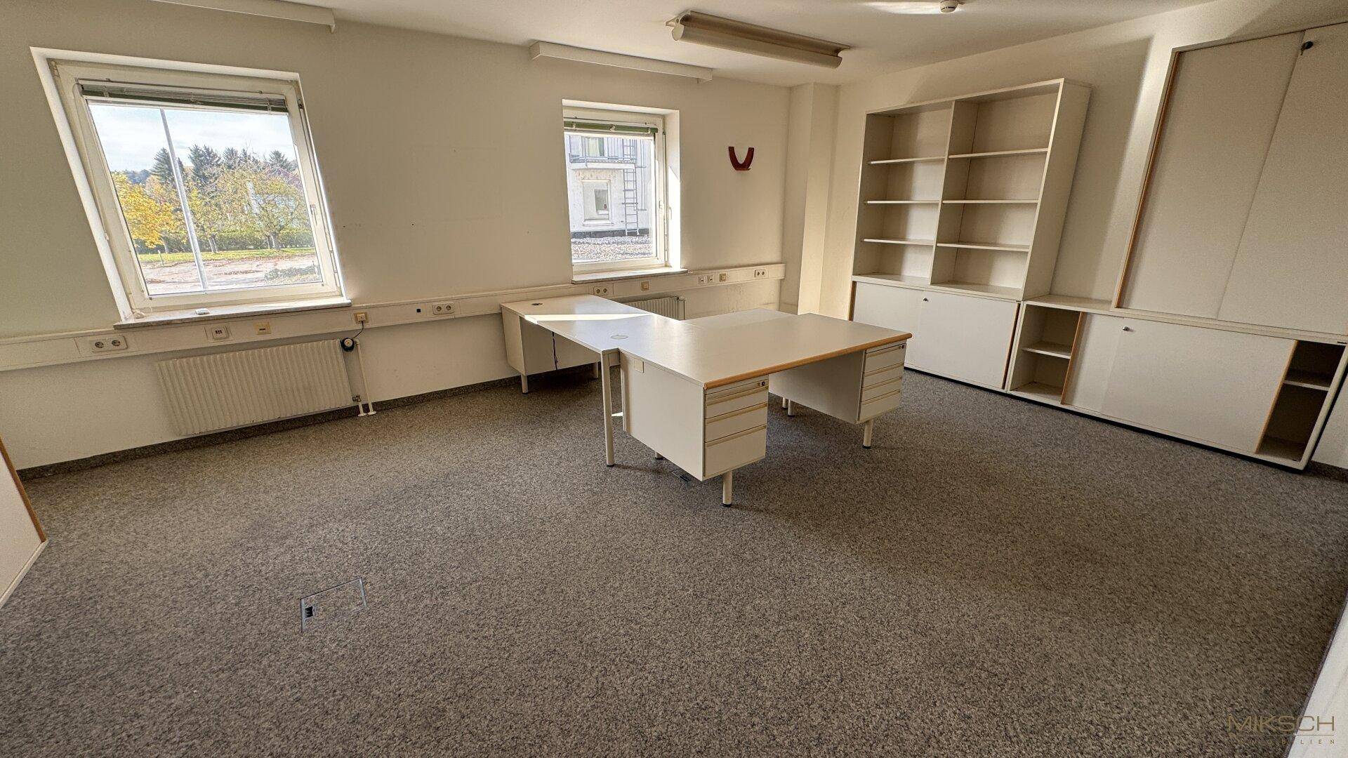 Helles Büro mit Teppichboden, weißen Schränken und einem großen Schreibtisch am Fenster.