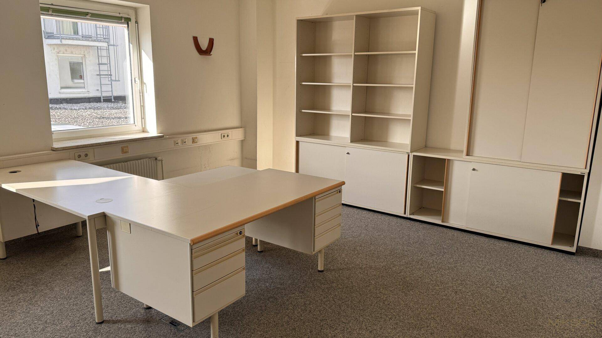 Büro mit Teppichboden, weißen Einbauschränken und einem Schreibtisch unter dem Fenster.