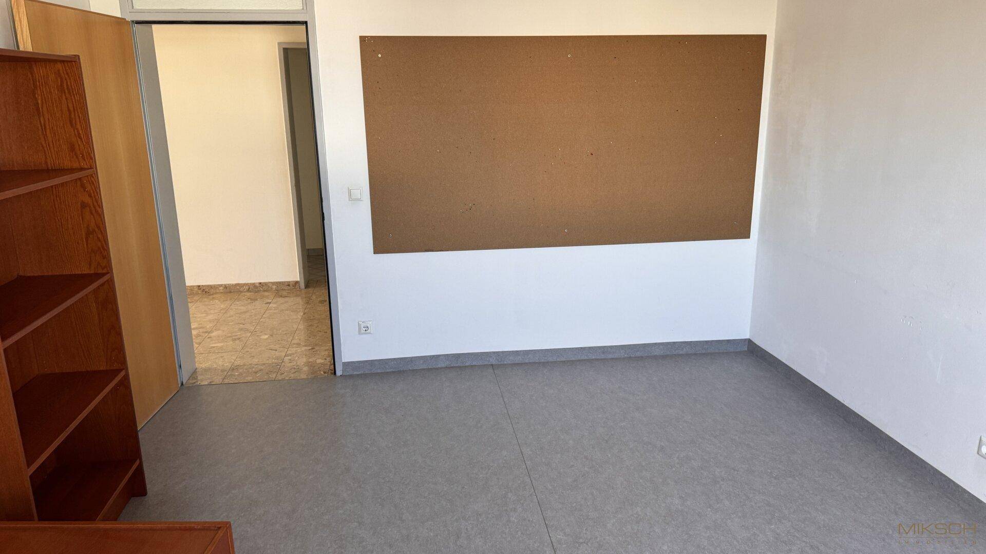Büro mit grauem Vinylboden, Pinnwand und Zugang zu einem weiteren Raum mit Fliesenboden.