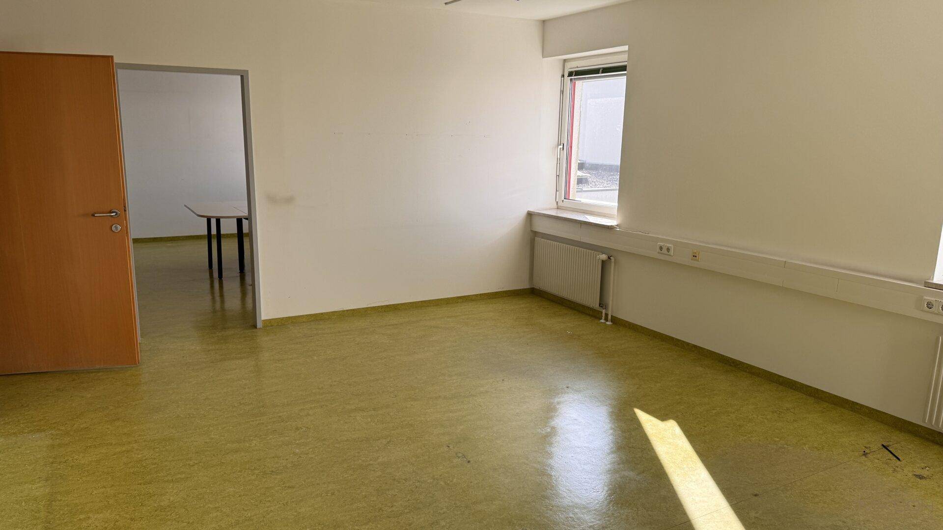 Büro mit grünem Linoleumboden, Fenster und Zugang zu einem weiteren Raum.