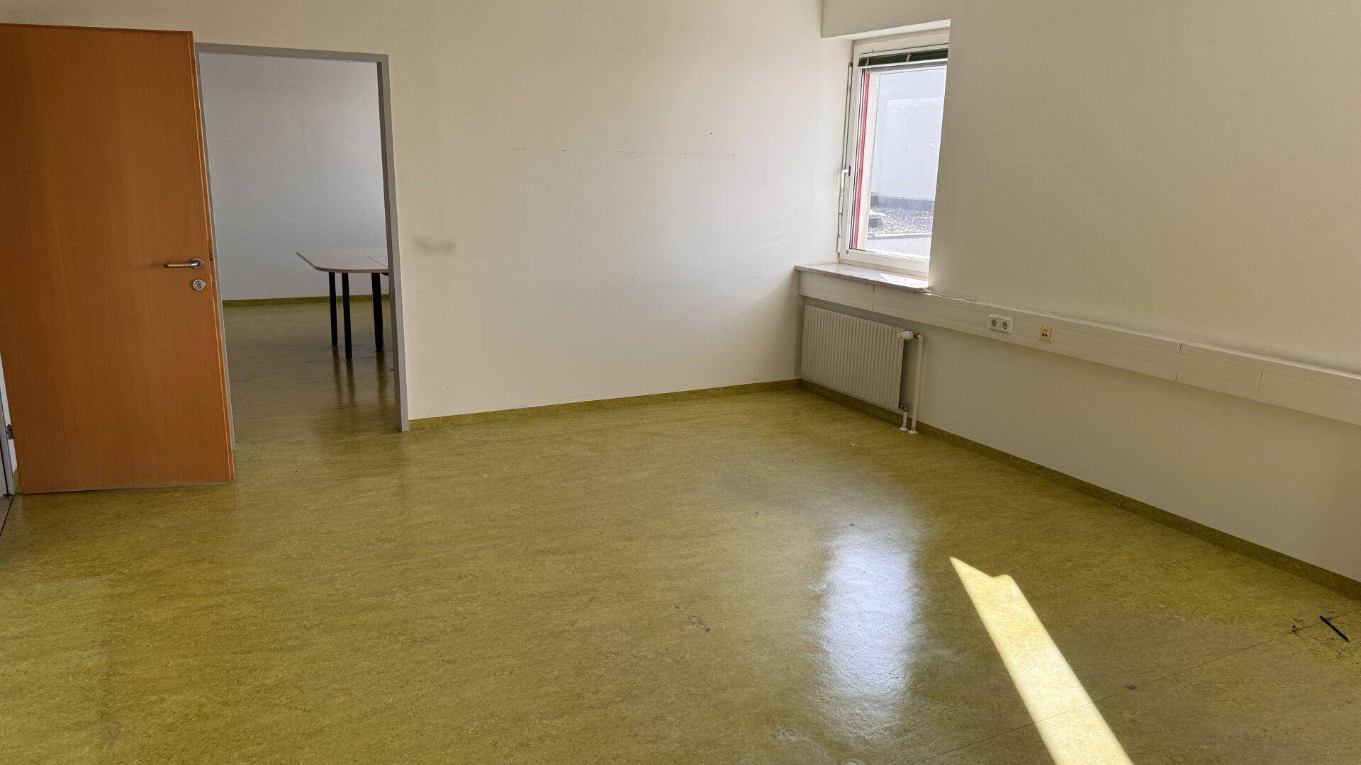 Büro mit grünem Linoleumboden, Fenster und Zugang zu einem weiteren Raum.
