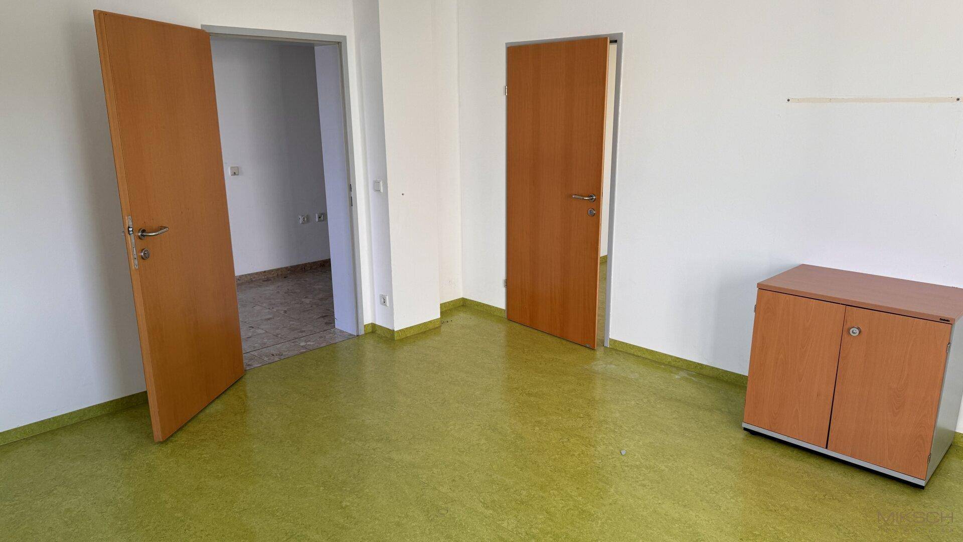 Büro mit grünem Linoleumboden und zwei Holztüren, die zu anderen Räumen führen.