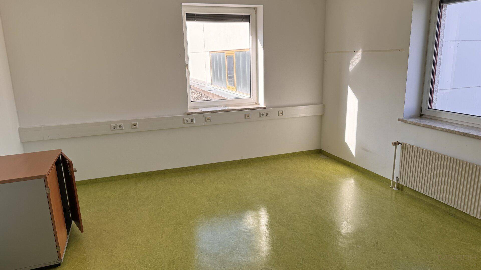 Helles Büro mit grünem Linoleumboden, zwei Fenstern und einem Heizkörper.