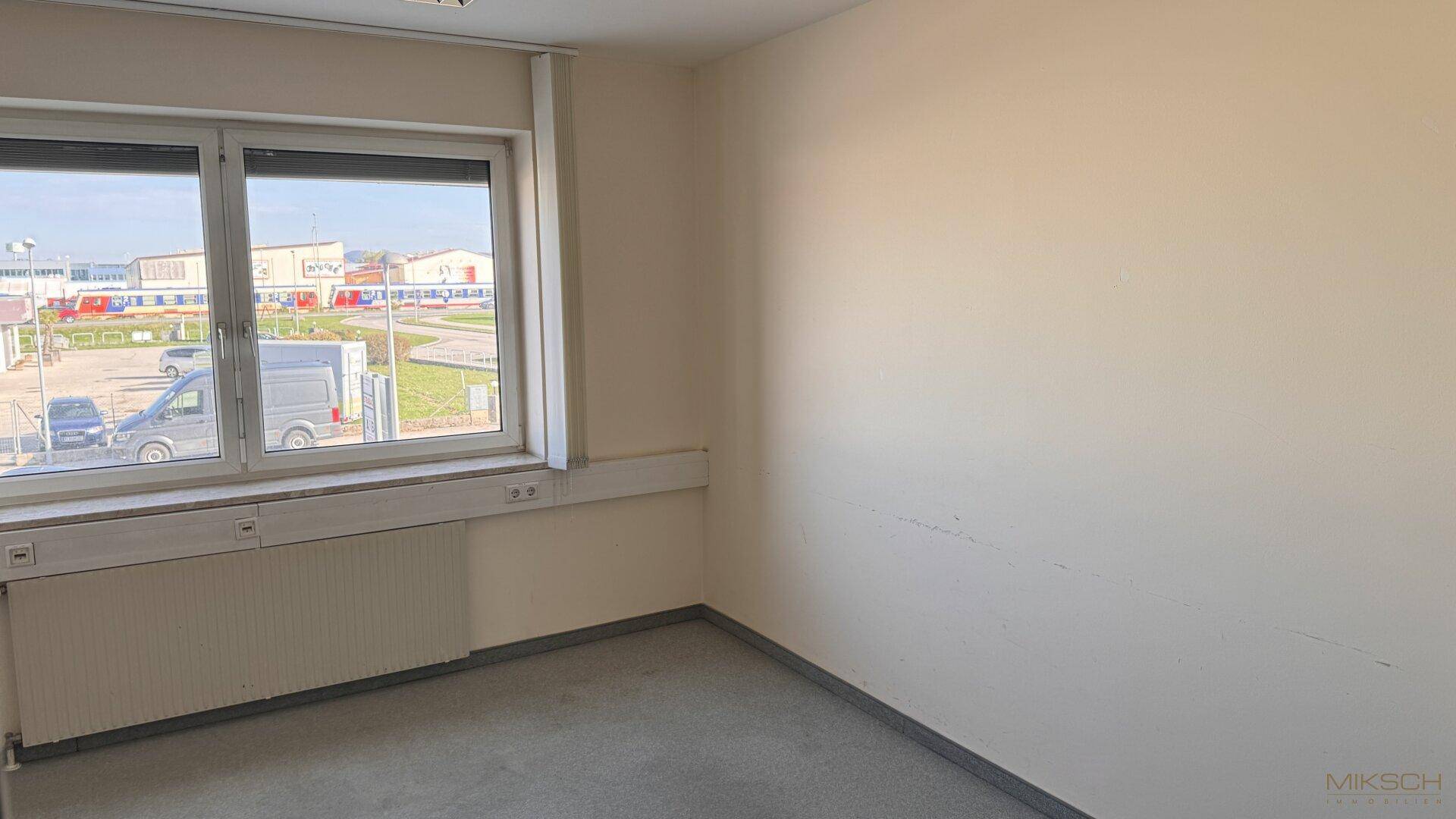 Büro mit grauem Teppichboden, großem Fenster und Blick auf ein Industriegebiet.