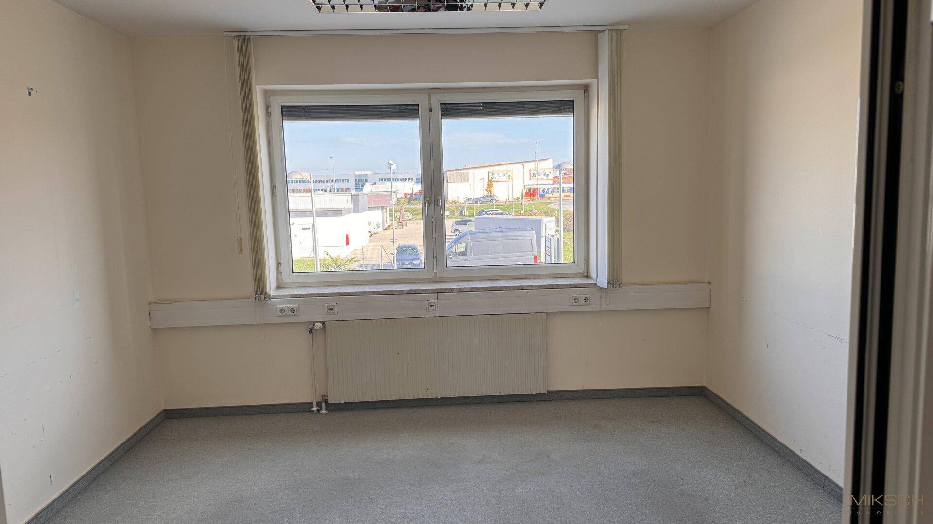Helles Büro mit grauem Teppichboden, großem Fenster und Blick auf die Umgebung.