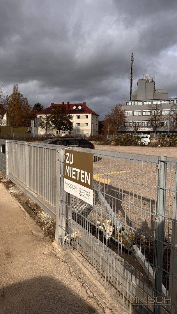 Nahaufnahme eines 'Zu vermieten'-Schildes an einem Zaun, mit Parkplatz und Gebäuden im Hintergrund.