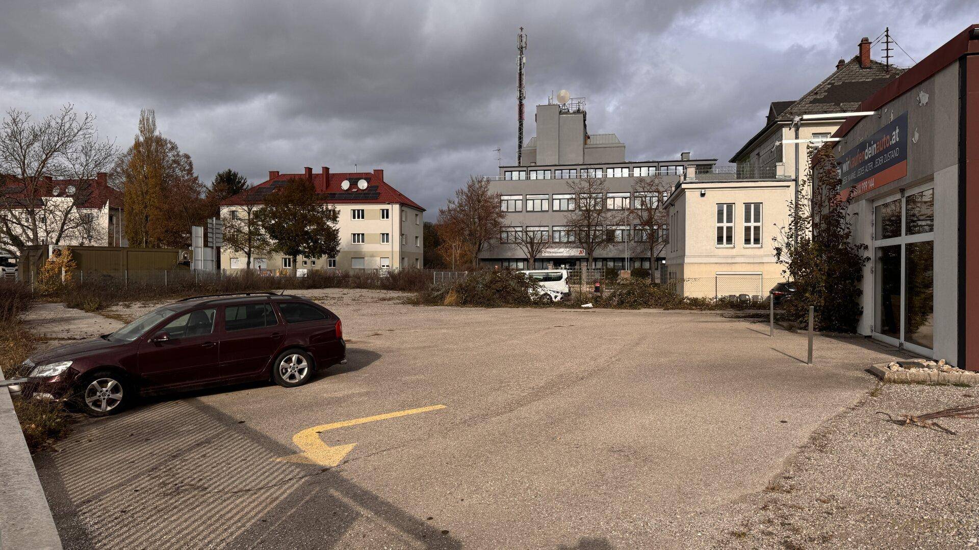 Befestigter Parkplatz mit einem geparkten Auto, umgeben von Gebäuden und Bäumen unter bewölktem Himmel.