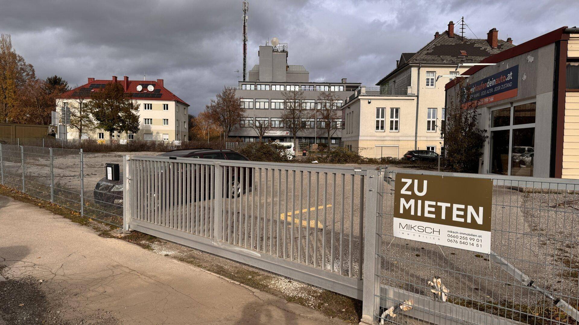 Eingezäunter Parkplatz mit 'Zu vermieten'-Schild, umgeben von Gebäuden und Bäumen unter bewölktem Himmel.