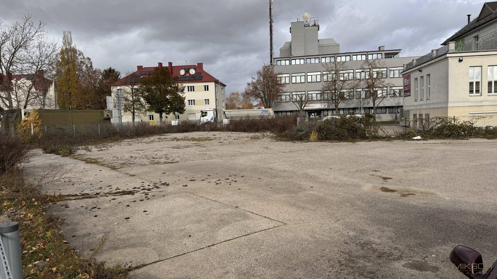 Weitläufiges, unbefestigtes Grundstück mit Schotter und umliegenden Gebäuden unter einem bewölkten Himmel.