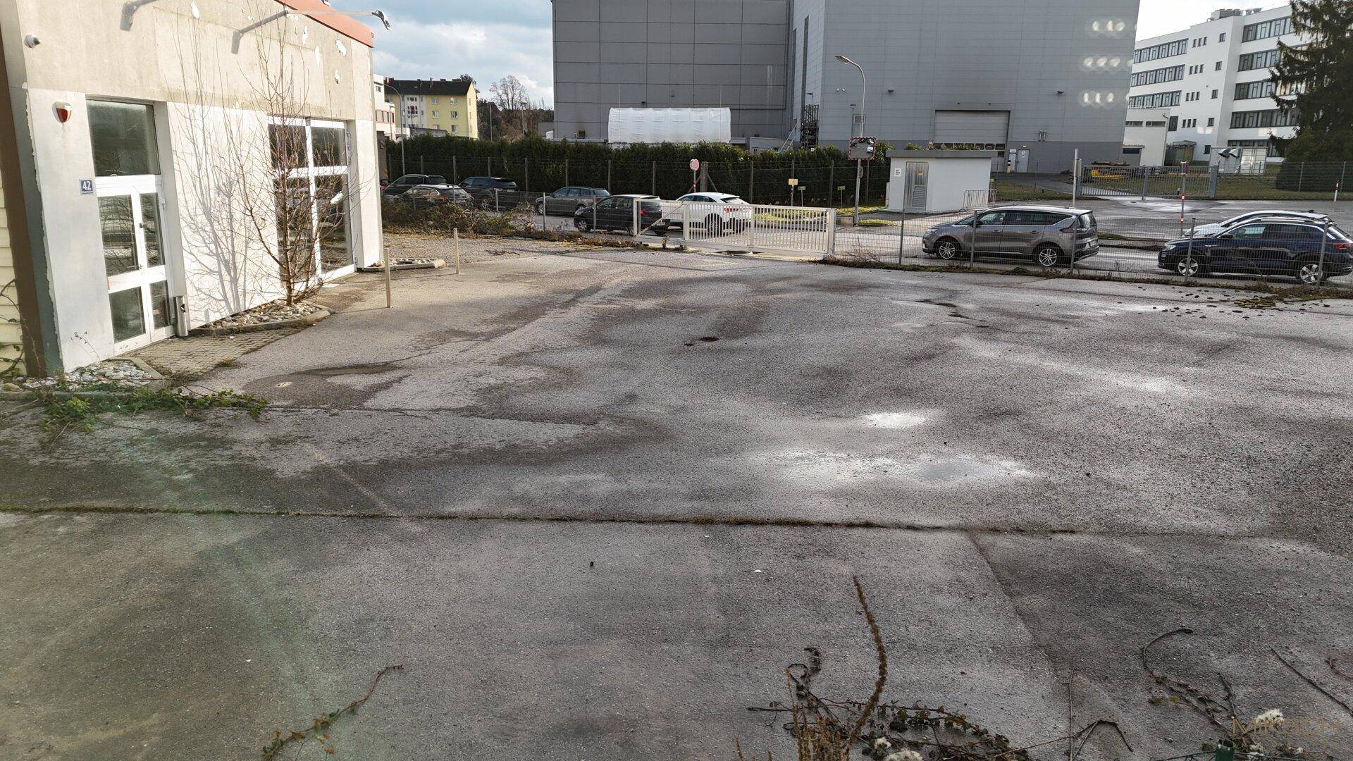Befestigter Parkplatz mit Spuren von Abnutzung neben einem Gebäude, umgeben von weiteren Geschäftsgebäuden.