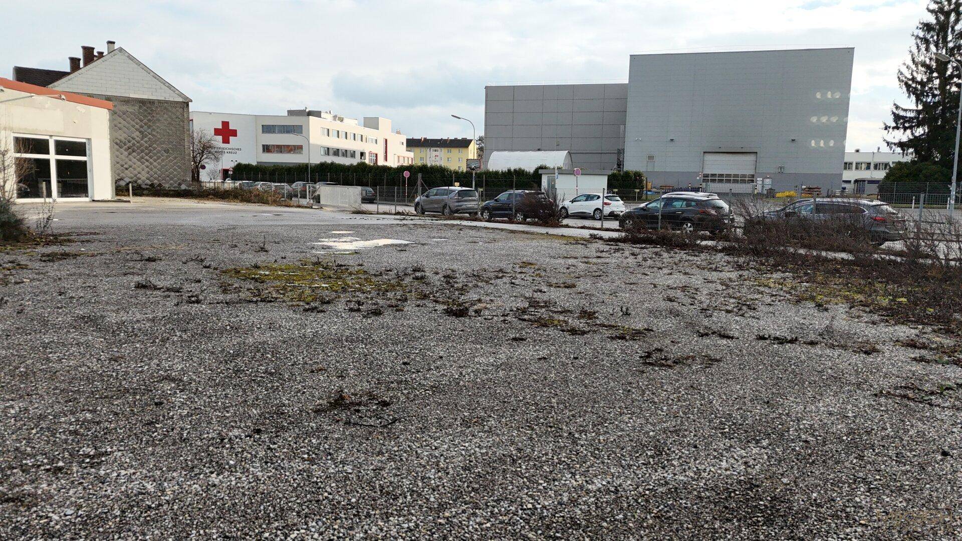 Großes, unbefestigtes Grundstück mit Schotterfläche, umgeben von Gebäuden und parkenden Autos.