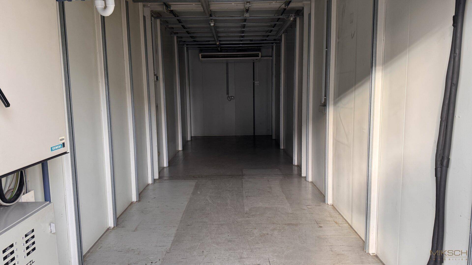Langer, schmaler Innenraum mit weißen Paneelwänden, Betonboden und sichtbarer Deckenkonstruktion.