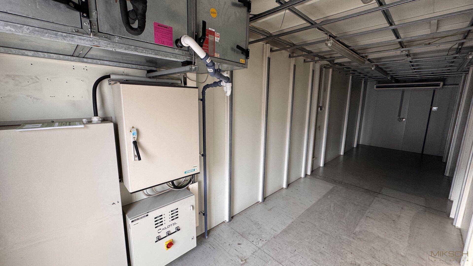 Technikraum mit Schaltschränken, Lüftungsanlagen und sichtbarer Deckenkonstruktion in einem langen Gang.