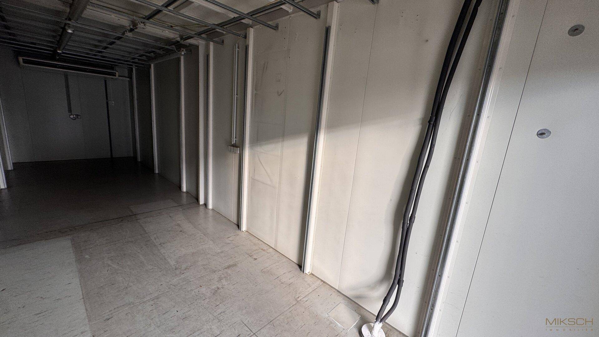 Schmaler Korridor mit weißen Paneelwänden, sichtbaren Kabeln und einem robusten Betonboden.