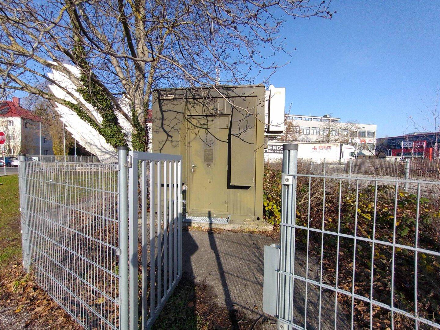 Grüner Technikschrank oder kleines Gebäude, eingezäunt, unter blauem Himmel mit Bäumen und Gebäuden im Hintergrund.