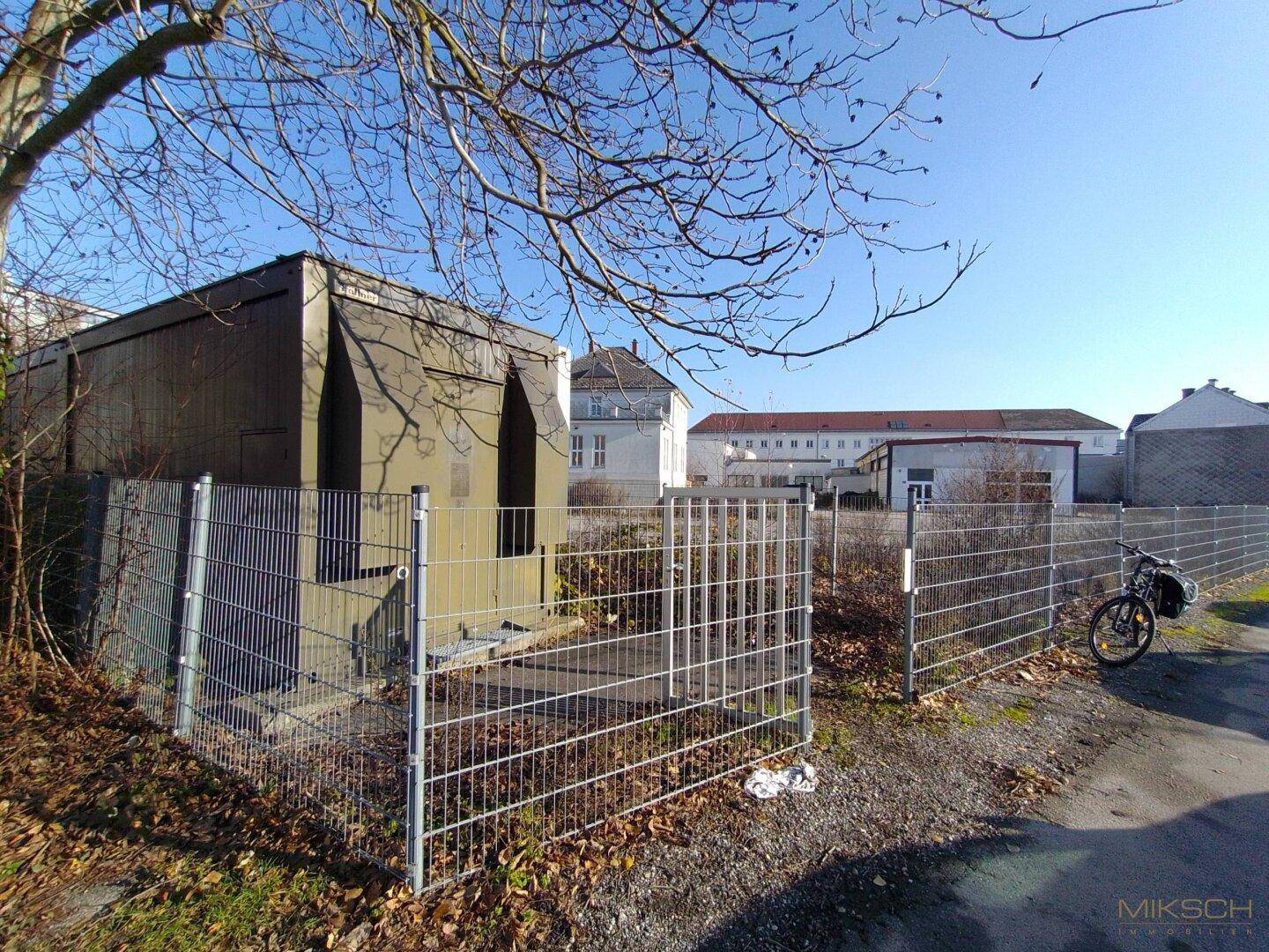 Grünes Technikgebäude, umgeben von einem Zaun, mit einem Fahrrad und weiteren Gebäuden im Hintergrund.