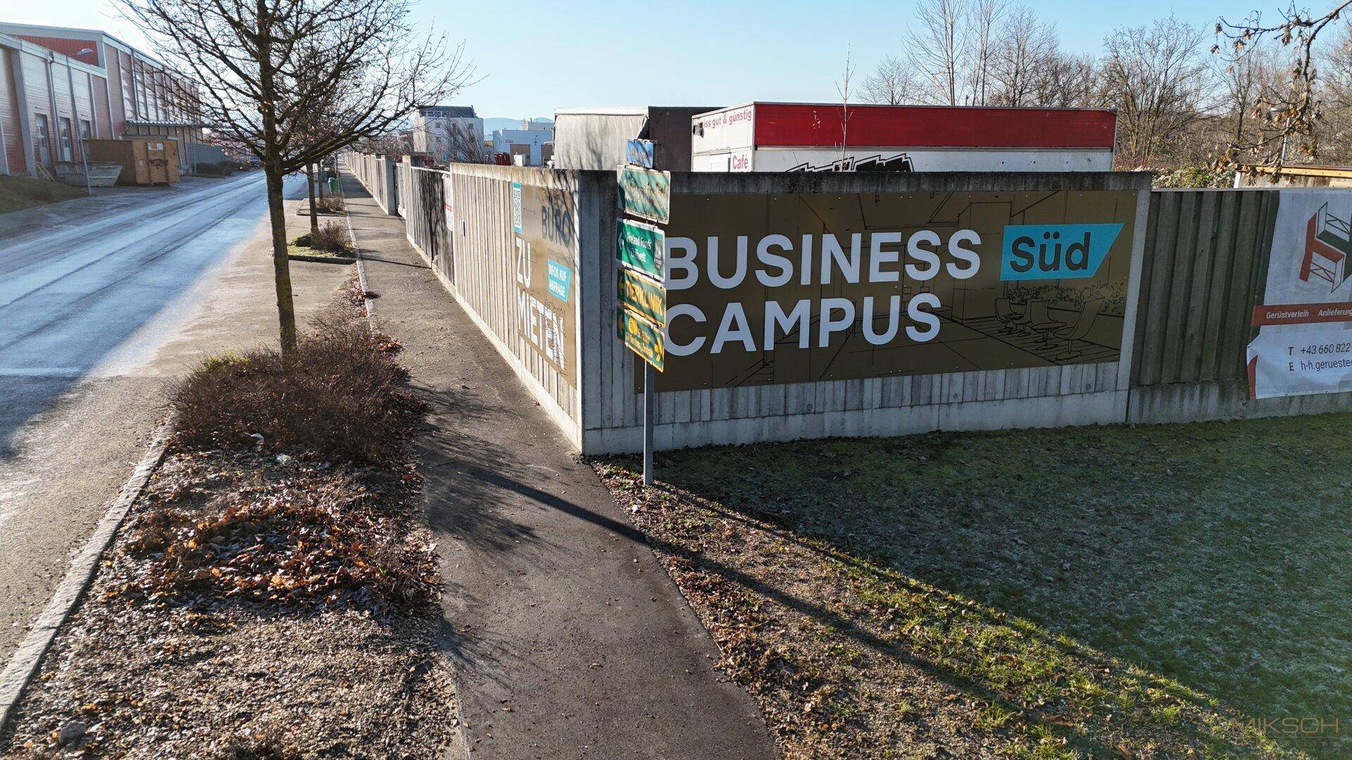 Eine lange Betonmauer mit einem "Business Süd Campus" Schild entlang eines Gehwegs.