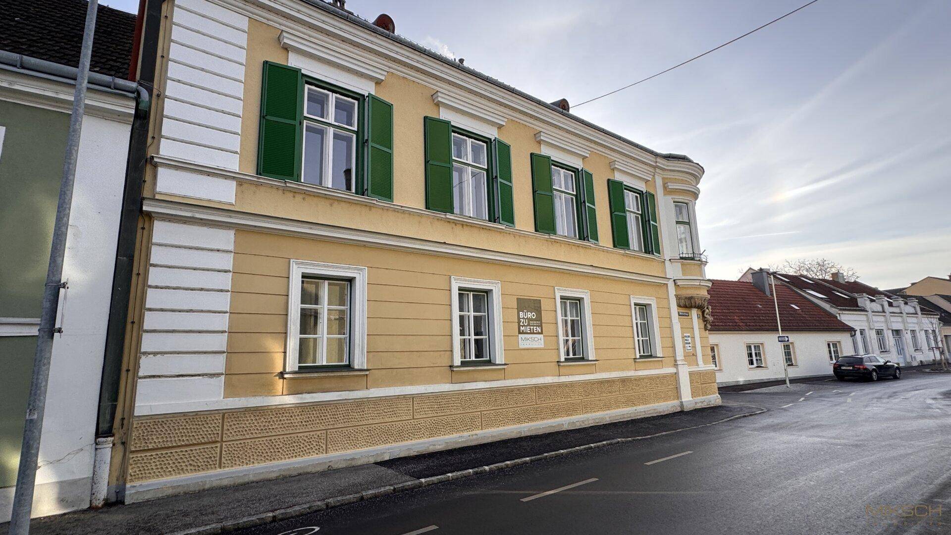 Seitenansicht eines historischen Gebäudes mit gelber Fassade, grünen Fensterläden und Blick auf die Straße.