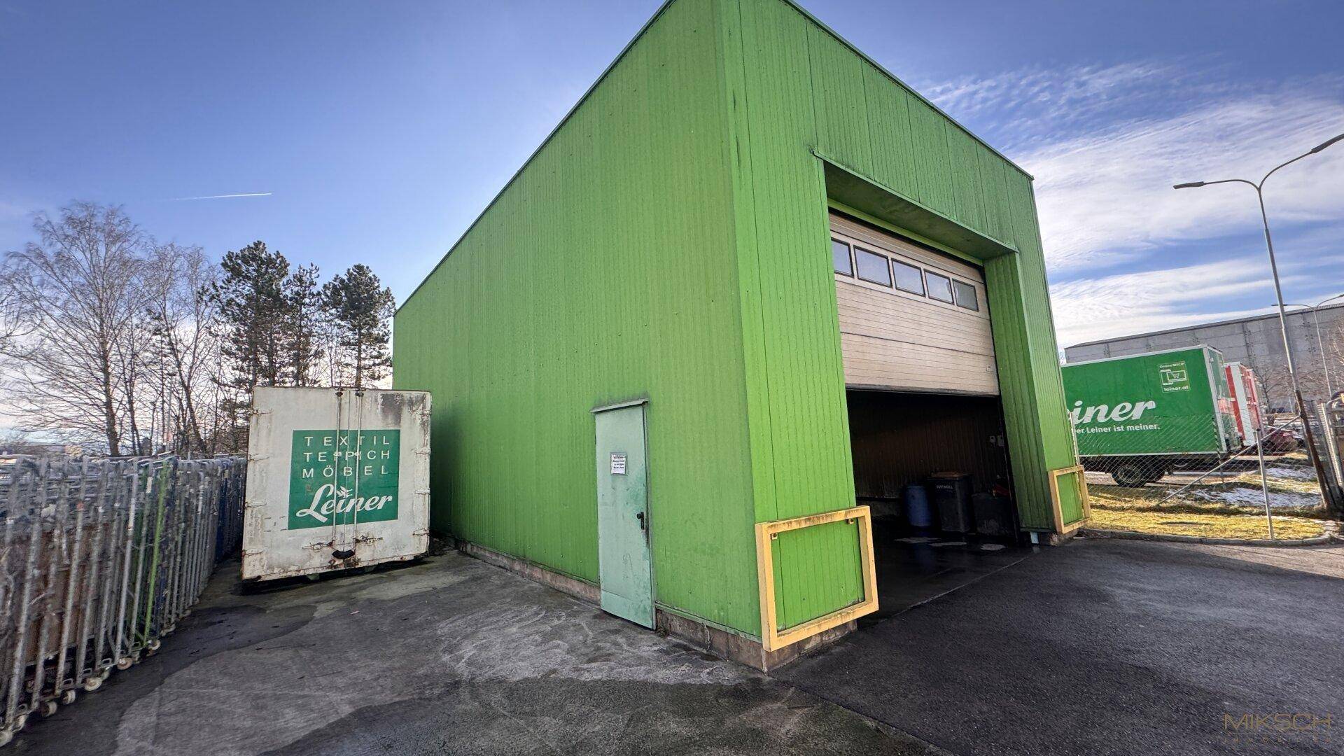 Grünes Industriegebäude mit großem Rolltor und kleinerer Zugangstür, typisch für Lagerhallen.