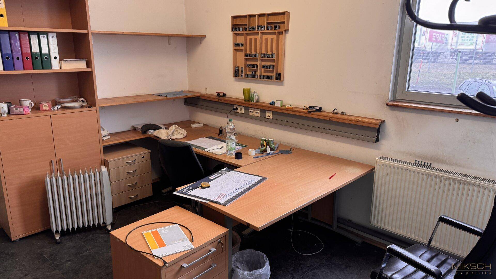 Büroarbeitsplatz mit Schreibtisch, Regalen und Heizkörper unter dem Fenster, ausgestattet für den täglichen Gebrauch.