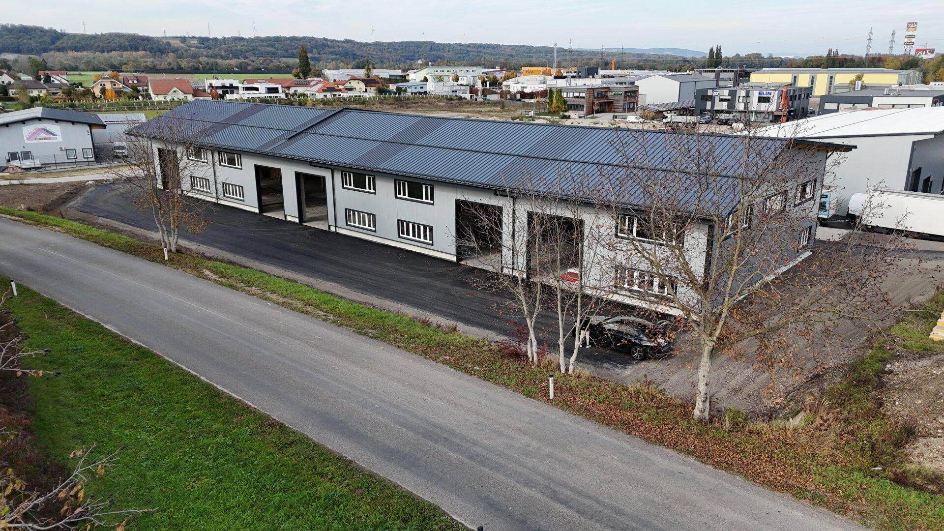 Weitläufige Luftaufnahme des modernen Industriegebäudes mit Zufahrtsstraße und ländlicher Kulisse.