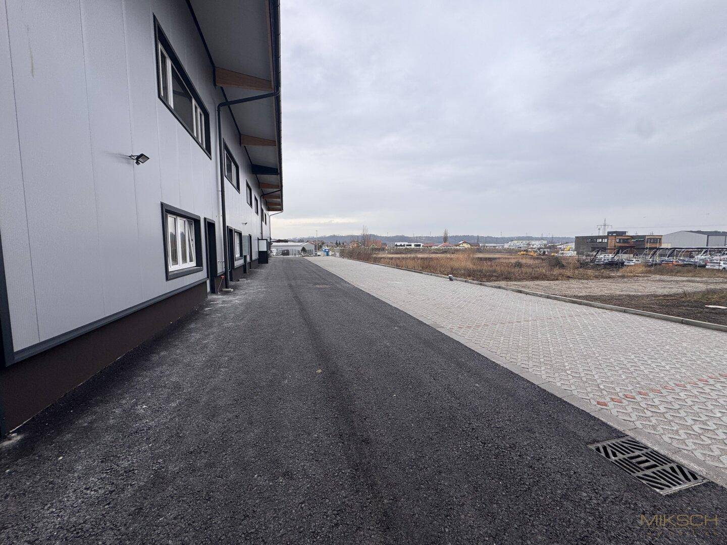 Seitenansicht der Gewerbehalle mit Zufahrtsweg und gepflastertem Bereich, unter bewölktem Himmel.