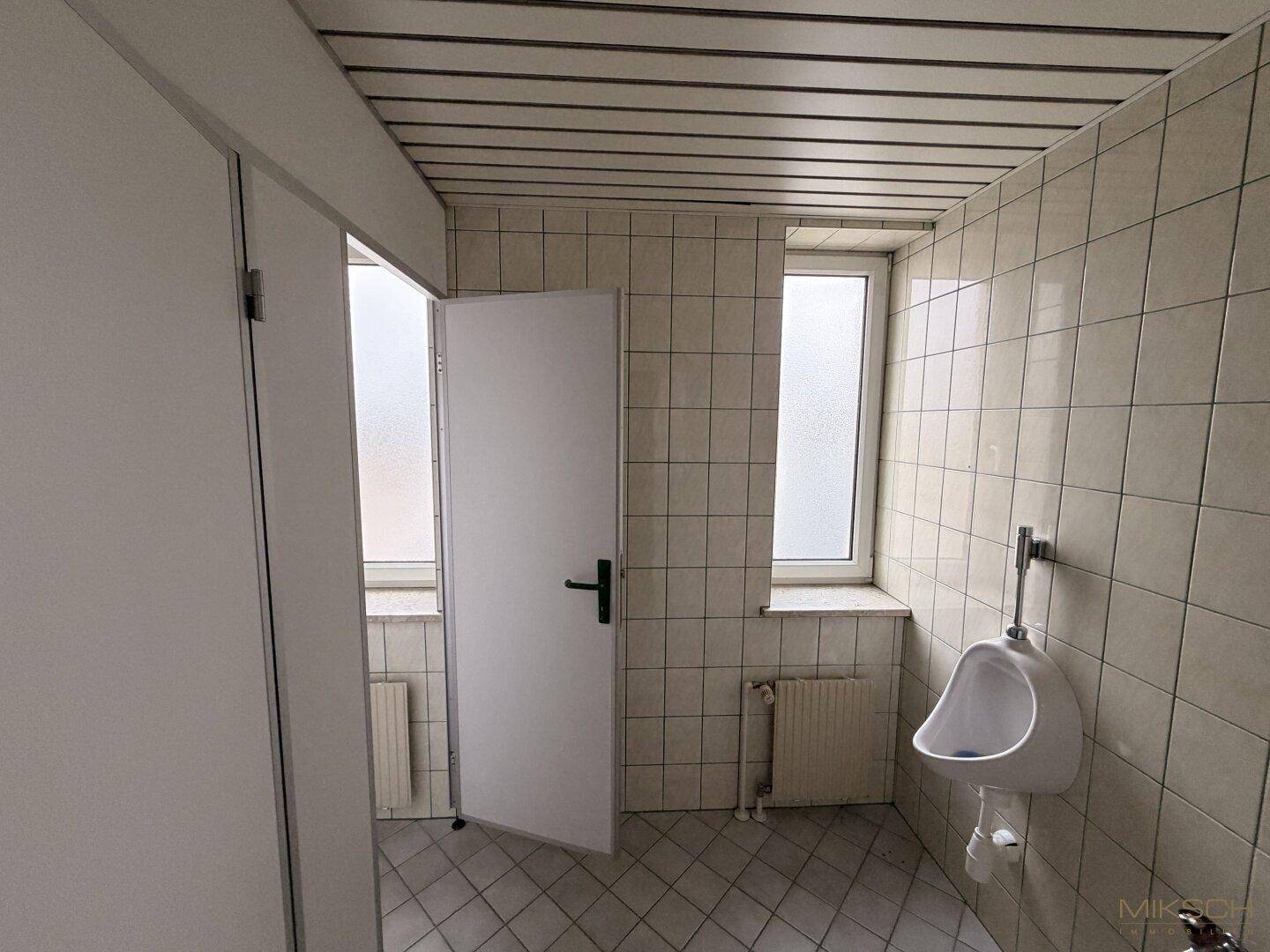 Herren-WC mit Urinal, hellbraunen Fliesen und einem Fenster für Belüftung.