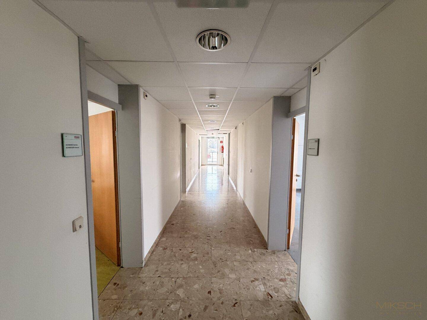 Langer Flur mit Fliesenboden, weißen Wänden und mehreren Türen zu Büroräumen.