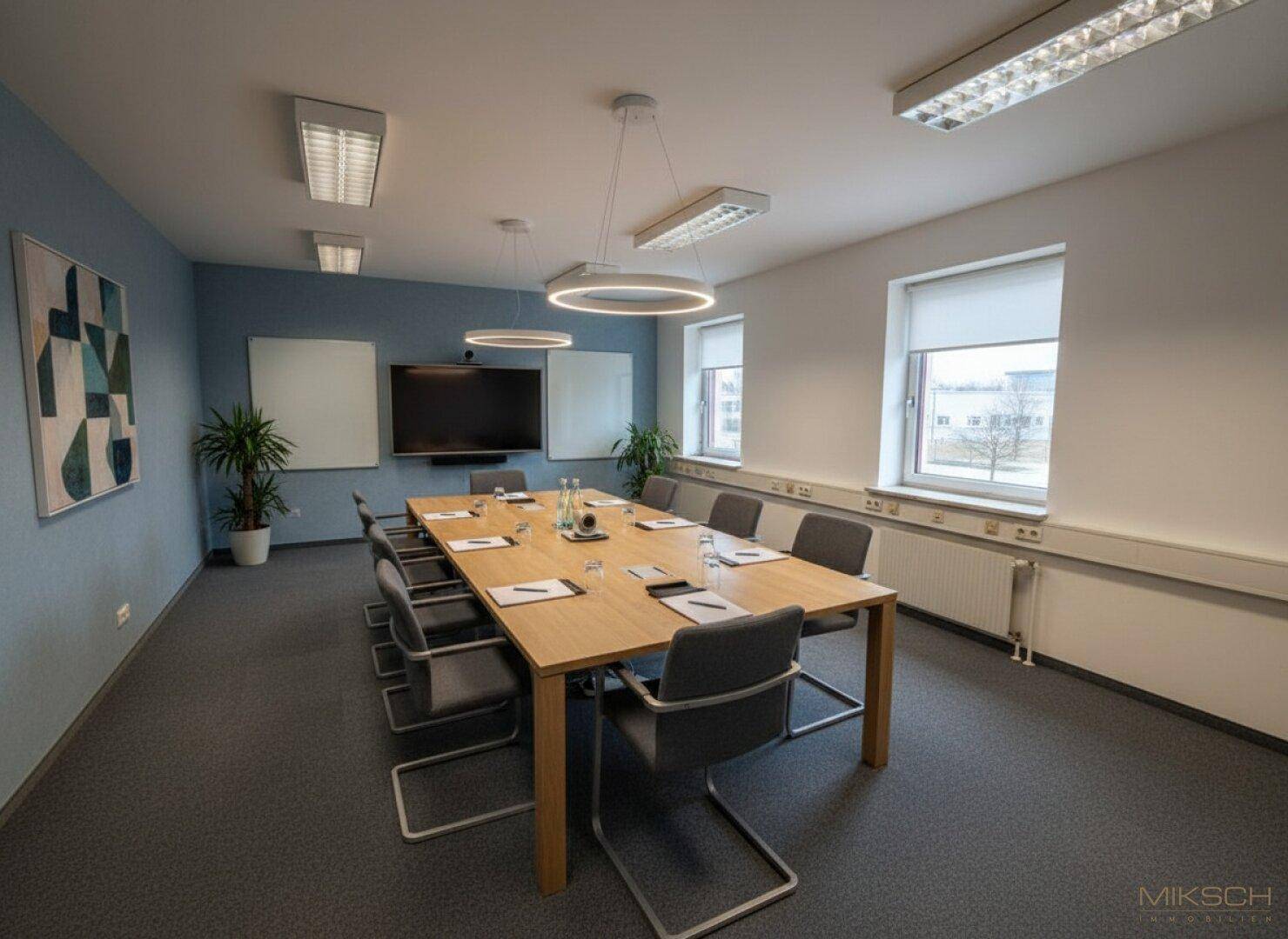 Moderner Konferenzraum mit großem Bildschirm, ovalen Deckenleuchten und Teppichboden.
