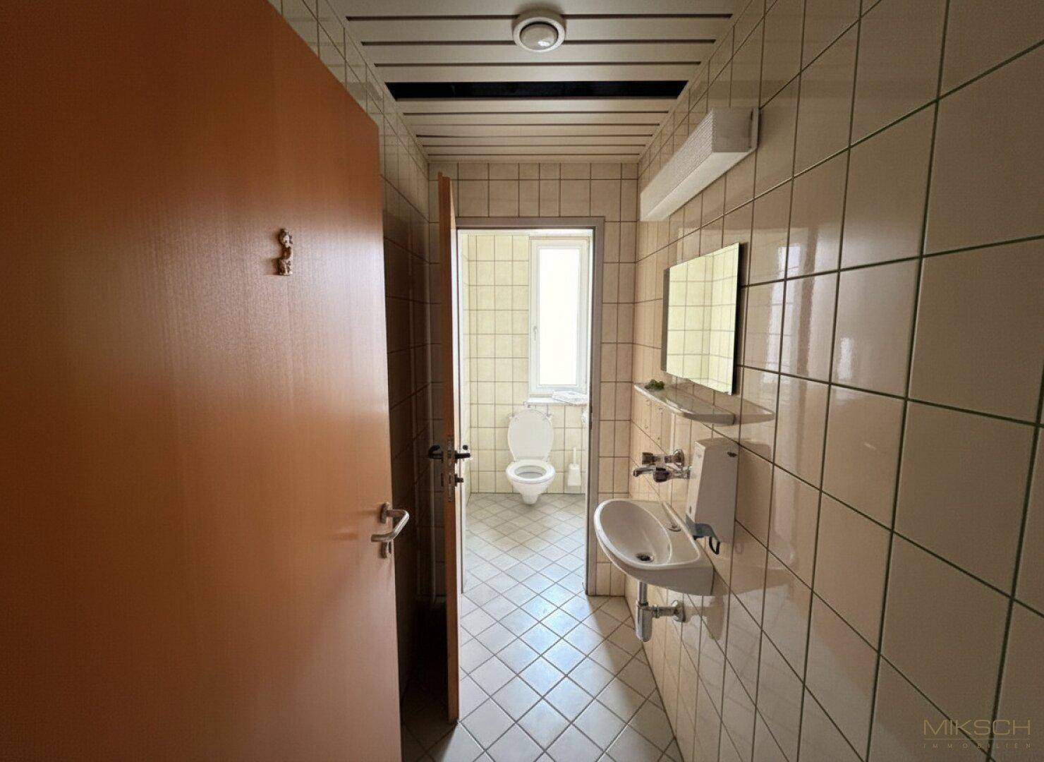 Toilettenraum mit hellbraunen Fliesen, Waschbecken und einer Toilette im Hintergrund.