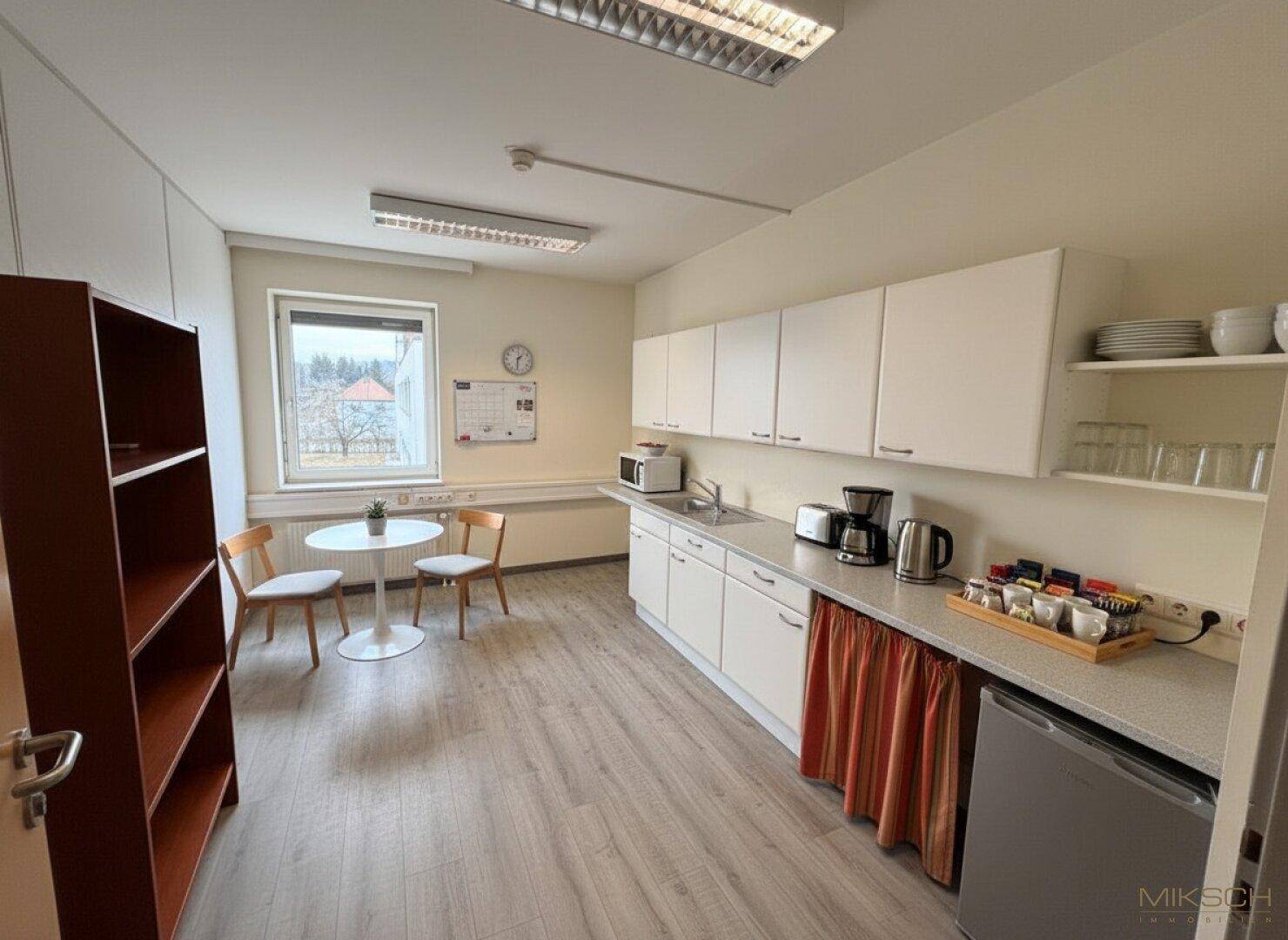 Kleine Teeküche mit weißen Schränken, Spüle, Mikrowelle und einem kleinen Tisch mit Stühlen.