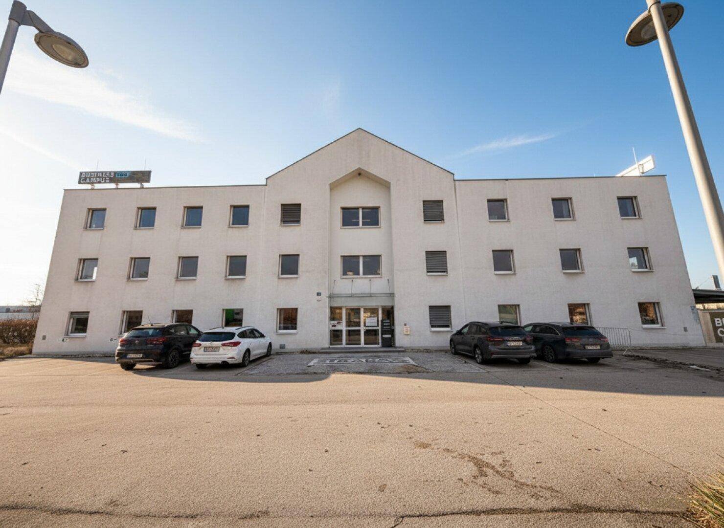 Modernes Geschäftsgebäude mit weißer Fassade, vielen Fenstern und Parkplätzen davor unter blauem Himmel.