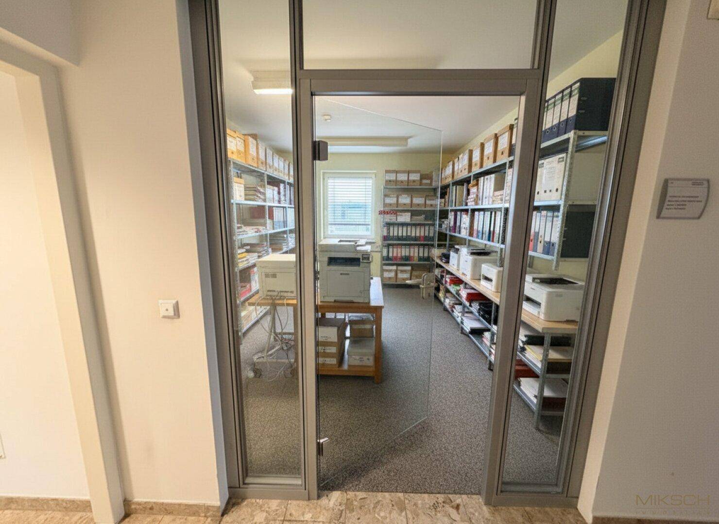 Archivraum mit Regalen voller Aktenordner, einem Fenster und einem Drucker in der Mitte.